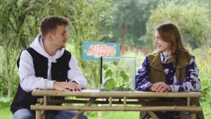 Een foto van de presentatoren Jurre en Emma die aan een bamboe tafel zitten en naar elkaar kijken. Op de tafel staat een glazen bak met takken en bladeren. Achter hen zijn struiken en het logo van het programma Steken en prikken.
