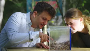 Een foto van de twee presentatoren aan een tafel. Ze zitten voorover gebogen te kijken naar een glazen bak met stro op de tafel. Jurre, aan de linkerkant, wijst naar de bak.