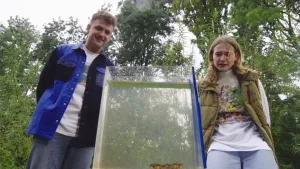 Een foto van de twee presentatoren die staan te kijken naar een smalle, hoge bak met water voor hen op de grond. Achter hen staan bomen.