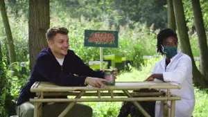 Een foto van twee mensen die aan tafel zitten. De presentator Jurre zit links en heeft een glazen pot vast die op tafel staat. Naast hem zit iemand in een doktersjas. Ze kijken allebei naar rechts. Achter hen staan struiken en het logo van het programma.