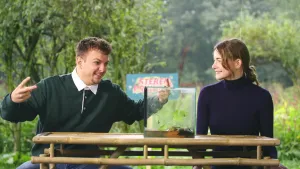 Een foto van de twee presentatoren, die aan een bamboe tafel zitten. Emma kijkt naar Jurre, die iets zit te vertellen. Jurre gebaart breed met zijn armen. Achter hen staan bomen en het logo van het programma Steken en prikken.