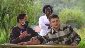 Een foto van drie mensen, van wie twee mensen aan een tafeltje zitten. Presentator Jurre heeft zijn onderarm plat op de tafel. Hij kijkt de andere kant op, terwijl de andere man tegen hem praat en een hand op zijn arm houdt. 