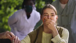 Een foto van presentator Emma met een hand voor haar mond. Bij haar andere arm is een paar handen van iemand anders te zien. Achter Emma staan twee mensen. Een van hen heeft een mondkapje op en staat voorovergebogen te kijken.