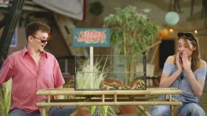 Een foto van presentatoren Jurre en Emma die aan een tafel zitten waar een bak met water op staat waar twee grote krabben inzitten. Presentator Emma houdt haar handen voor haar mond terwijl ze naar de bak kijkt.