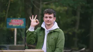 Een foto van presentator Jurre die aan een tafel zit. Hij houdt een hand omhoog met een oké-teken: met zijn duim en wijsvinger vormt hij een rondje en drie vingers staan omhoog. Achter hem staat bomen en een bordje met het logo van het programma.