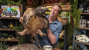 Een foto van presentator Freek die in de studio staat en een alligatorschildpad in zijn handen heeft en die laat zien. De schildpad heeft een open bek en kijkt richting Freek.