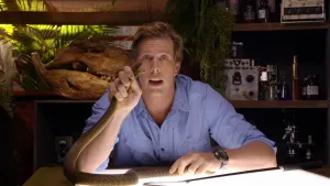 Een foto van presentator Freek die in de studio aan een lichtgevende tafel zit en de kop van een slang in zijn hand heeft. De slang heeft een geopende bek.