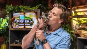 Een foto van presentator Freek met een jonge kat in zijn handen. Hij geeft de kat een kusje op de wang. Achter hem zijn veel planten te zien en het logo van Freeks Jungle School.