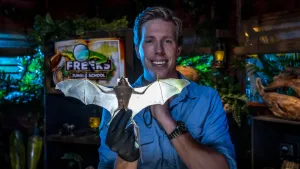 Een foto van presentator Freek met een opgezette vleermuis in zijn handen. De vleugels van de vleermuis zijn helemaal uitgerekt. De kamer waarin Freek staat is best donker en op de achtergrond is het logo van Freeks Jungle School te zien.