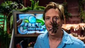 Een foto van presentator Freek met een vogelspin op zijn blije gezicht. Achter hem staat een beeldscherm met het logo van Freeks Jungle School. Er is ook een deel van een krokodillenhoofd te zien en veel planten.