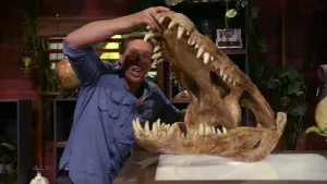 Een foto van presentator Freek met in zijn handen de schedel van een krokodil. De kaak van de krokodil is wijd open, van Freeks borstkas tot boven zijn hoofd. Freek staat naast een hoge tafel.