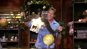 Een foto van presentator Freek met in zijn handen een schedel van een buffel. De witte schedel heeft twee grote donkere hoorns naar opzij. Achter hem staan kasten vol spullen en planten. 