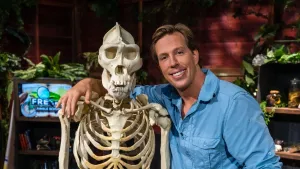 Een foto van presentator Freek die naast het skelet van een gorilla staat. Hij heeft zijn arm om de schouders van het skelet. Links achter hem is het logo van het Freeks Jungle School te zien.