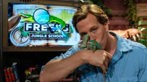 Een foto van presentator Freek met een kameleon op zijn hand. Hij geeft de kameleon een kusje op zijn rug. Achter hem op de muur is het logo van Freeks Jungle School te zien.