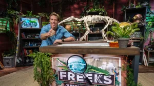 Een foto van presentator Freek achter een tafel met daarop een skelet van een reuzenmiereneter. Achter hem in de kamer staan kasten vol spullen en veel planten. Voor op de tafel is het logo van Freeks Jungle School te zien.