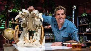 Een foto van presentator Freek met naast hem op tafel een schedel van een nijlpaard. Hij tilt de kaak open zodat alle tanden goed te zien zijn. Achter hem staan kasten vol met spullen en planten.