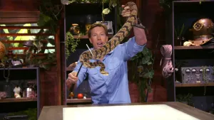 Een foto van presentator Freek met een grote slang in zijn handen. Met een hand gebruikt hij een stok met haak om de slang vast te houden. Hij staat achter een tafel met een groot wit vlak. Achter hem staan kasten en planten.