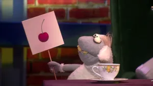 Een foto van pop Stella Schaap. De pop heeft een bord met één getekende roze kers erop vast en kijkt naar het bord. De pop heeft een witte vacht op haar rug en oren, een grijze vacht bij kop en poten, ronde ogen en vooruitstekende, gele tanden.