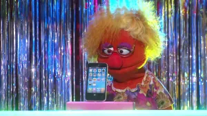 Een foto van een pop die Jannie heet en kijkt naar een rechthoekig roze blok met een mobiele telefoon erop, een Apple iPhone. De pop heeft een rode huid, lichtbruin haar, een bril op en een ronde neus. De pop draagt een jurk met bloemetjes.