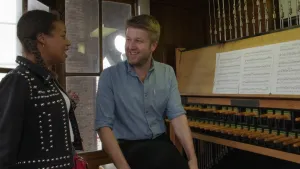 Een foto van Eva die naast beiaardier Richard staat, die op een carillon kan spelen. Dat is een soort piano met houten ronde staven als toetsen. Erboven zitten stalen kabels, waarmee de staven vastzitten aan de niet-zichtbare klepels van klokken.