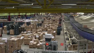 Een foto van de grootste opslagplaats van boeken die in Nederland verkocht worden. Er staan stapels kartonnen dozen vol met boeken op rekken en houten laadborden. Rechts zijn een zwarte sorteerband en stalen vakken te zien met kartonnen dozen erop.