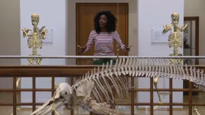 Een foto van Sosha in een gang. Links en rechts van Sosha is een animatie van een menselijk skelet te zien die dansen met gebogen benen en gekruiste handen. Ervoor hangt een skelet van een dier met een lange bek, twee grote voorpoten en een lange rug.