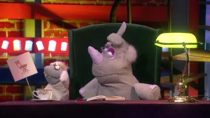 Een foto van pop Stelle Schaap met een bord waarop een getekende, bruine klapstoel staat. De pop Nico de Neushoorn zit op een groene stoel met een bril op. Voor Nico op tafel liggen een notitieboek en pen en staan een kopje thee en een bureaulamp.