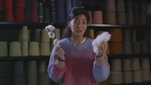 Een foto van een presentator. Ze kijkt in de camera en houdt een takje van een katoenplant vast. Achter haar zie je planken die vol staan met rollen met garen, in allerlei kleuren.