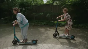 Een foto van twee mensen, die allebei op een elektrische step rondrijden. Ze rijden op een stuk plein, met daarachter bomen en een schutting.