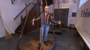 Een foto van presentator Janouk in een kamer. Janouk staat op een van twee oude houten platen. De platen hangen aan elk vier touwen. Aan de rechterkant van de kamer is een lange bank. Achter Janouk staat een trap, en een bezem tegen de muur.