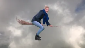 Een foto van presentator Janouk, die op een bezemsteel vliegt. Achter haar zijn wolken te zien.