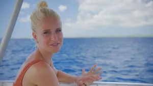 Een foto van presentator Janouk. Ze zit op een boot op zee, en praat tegen de kijker. De zee is blauw van kleur. 