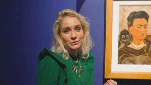 Een foto van presentator Janouk. Ze praat in de camera. Achter haar hangt een schilderij aan een blauwe muur. Op dat schilderij is de kunstenaar Frida Kahlo te zien.
