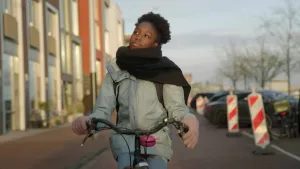 Een foto van een meisje op een fiets. Ze is ongeveer veertien jaar. Ze fietst in een straat, richting de kijker. Ze kijkt schuin omhoog naar iets buiten beeld. 
