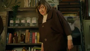 Een foto van een vrouw. Ze is warm gekleed. Ze staat in een kamer met een oude inrichting en kijkt naar benden.  In een kast achter haar zie je oude boeken en glazen potten. Er hangt ook een poster van een oude man met een bolhoed en een witte baard.