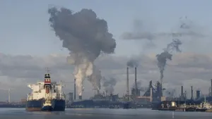 Een foto van industrie aan het water. Je ziet fabrieken met schoorstenen waar grote rookwolken uit komen. Links in beeld zie je een groot schip dat gesleept wordt door een sleepboot. 