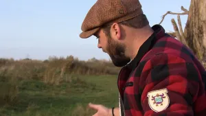 Een foto van boswachter Tim in de natuur. Hij heeft een pet op en zijn bekende rood-zwart-geblokte jas aan. Hij kijkt naar iets op de grond.