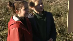 Een foto van twee jonge meisjes in de natuur. Ze kijken naar een bord waarvan je een stukje van de achterkant ziet. Ze glimlachen. 