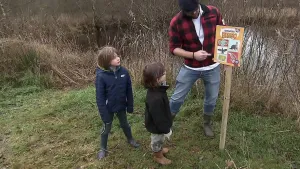 Een foto van twee jonge jongens en de presentator, in de natuur. Ze kijken alle drie naar een bord op een houten paal. Daarop staat de beestenbrigade-bingo. Achter hen zie je een sloot.