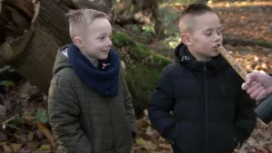 Een foto van twee jonge jongens in een bos. Een van hen praat tegen de onderkant van de steel van een hamer, alsof het een microfoon is. 