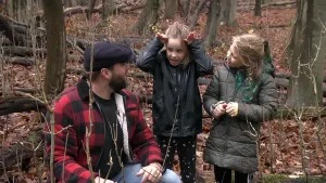 Een foto van presentator Tim die in het bos op de grond zit en met twee meisjes aan het praten is. Een van de meisjes houdt haar handen naast haar hoofd alsof ze een hert nadoet.