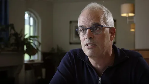 Een foto van een man in een kamer. Hij heeft grijs haar en een bril op. Hij kijkt naar iets naast de camera, en is wat aan het vertellen.