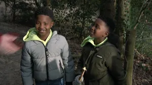 Een foto van twee jonge jongens in de natuur. Ze hebben winterjassen aan. Een van hen houdt een stok vast. Links in beeld zie je nog net een bewegende hand. 