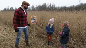 Een foto van presentator boswachter Tim die met twee kleine meisjes naast het riet staan. Tussen hun in staat er een houten paal met een beestenbrigade bingo kaart erop.