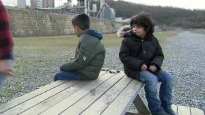 Een foto van twee jongentjes die buiten op een tafel zitten met hun ruggen naar elkaar toe. Op de achtergrond is er een industrie bedrijf te zien.