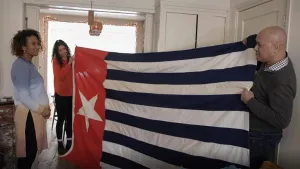 Een foto van twee mensen die in een kamer een heel grote vlag van Nederlands-Nieuw-Guinea vasthouden en die laten zien aan een vrouw die naast hun staat toe te kijken.