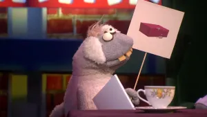 Een foto van pop Stella Schaap met een witte vacht die grijs is bij kop en poten, met ronde ogen en vooruitstekende, gele tanden. De pop heeft een bord vast waar een getekende rode baksteen met enkele spetters grijs cement erop te zien is. 