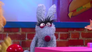 Een foto van pop Peter Allesweter achter een roze tafel. De pop heeft een lichtblauwe vacht, een paarse ronde neus en grote, ronde ogen met lange, zwarte wimpers. De pop heeft lange rechtopstaande oren, zoals een konijn of haas. 