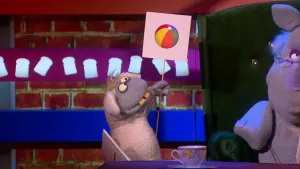 Een foto van pop Stella Schaap met vooruitstekende, gele tanden. De pop heeft een bord vast met een getekende strandbal met gele groene, rode en blauwe vlakken erop en kijkt ernaar. De pop staat naast de groene stoel van pop Nico de Neushoorn. 