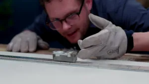 Een foto van een persoon die over een tafel leunt en kijkt naar een klein metalen blokje op een lange metalen lat of rails. Hij heeft dikke handschoenen aan. Er komt wat witte damp van de tafel af.